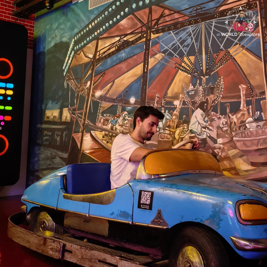 Her husband sitting in an old blue bumper car in front of a funfair carousel mural, at Neonia