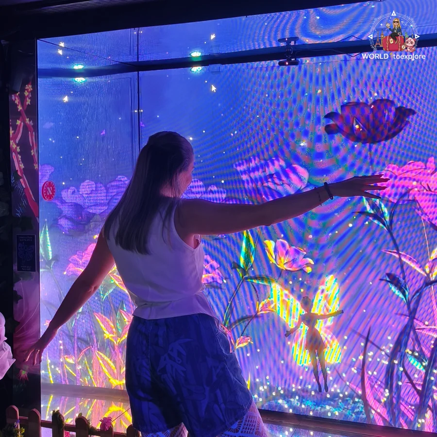 Cristina with her arms open beside an interactive screen with colourful flowers and a fairy, in a room at Neonia