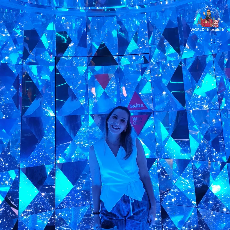 Cristina smiling in a mirror room with blue triangles, next to an exit sign, at Neonia