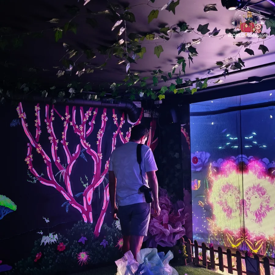Her husband with a backpack looking at a forest scene with pink neon branches, in a dark room at Neonia