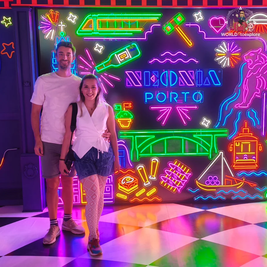 Cristina and her husband hugging in front of the Neonia Porto neon panel, with the bridge, the tram and the rabelo boat drawn in coloured lights