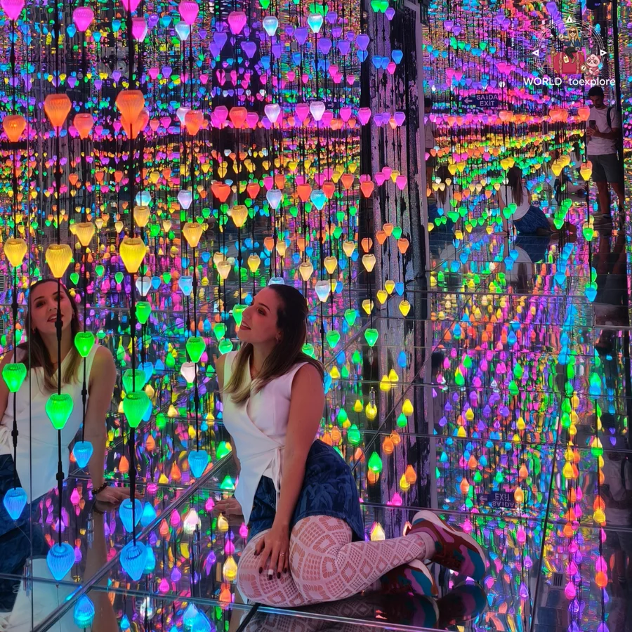 Cristina kneeling on the floor of a mirror room full of coloured lights, looking up, at Neonia