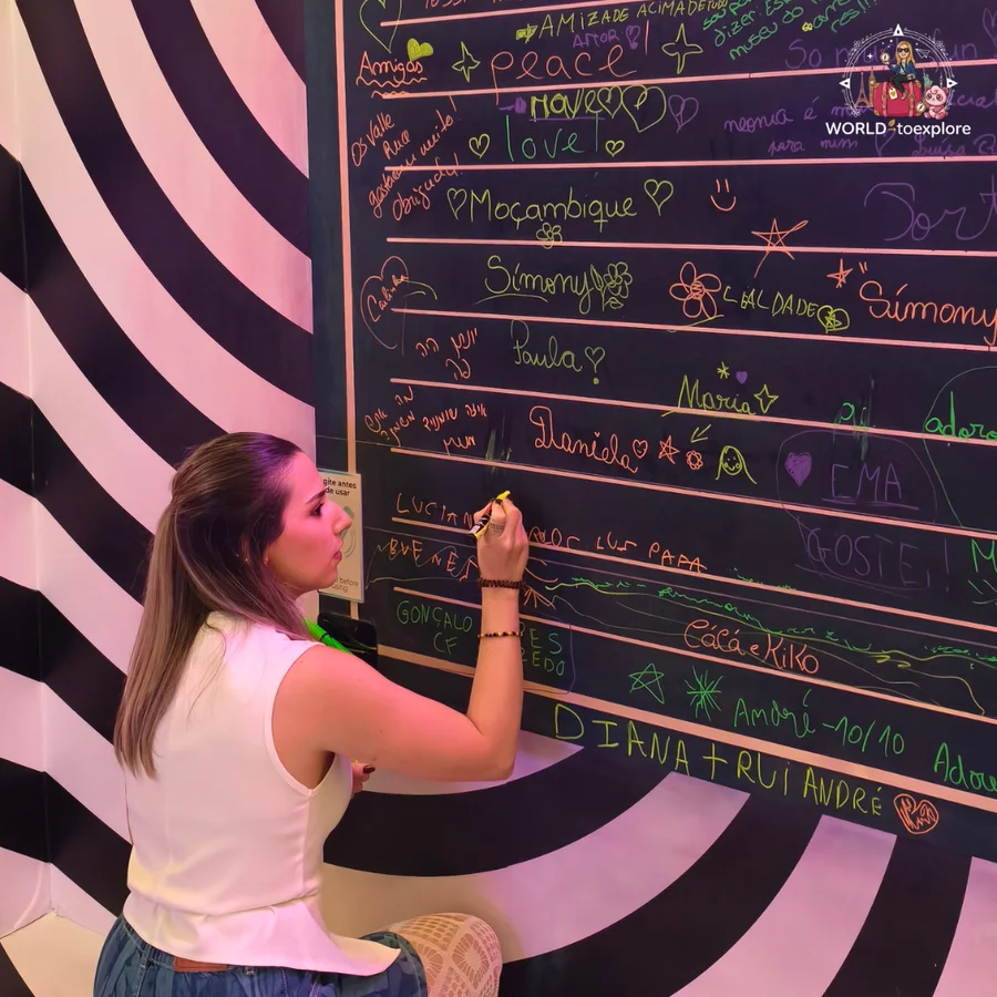 Cristina writing in chalk on a blackboard full of visitors' messages, beside a black-and-white striped wall, at Neonia