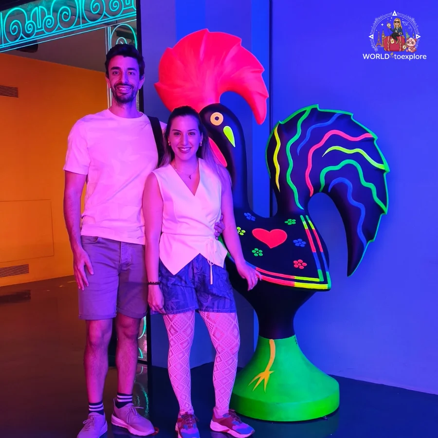 Cristina and her husband beside a giant, colourful Barcelos rooster in a room at Neonia