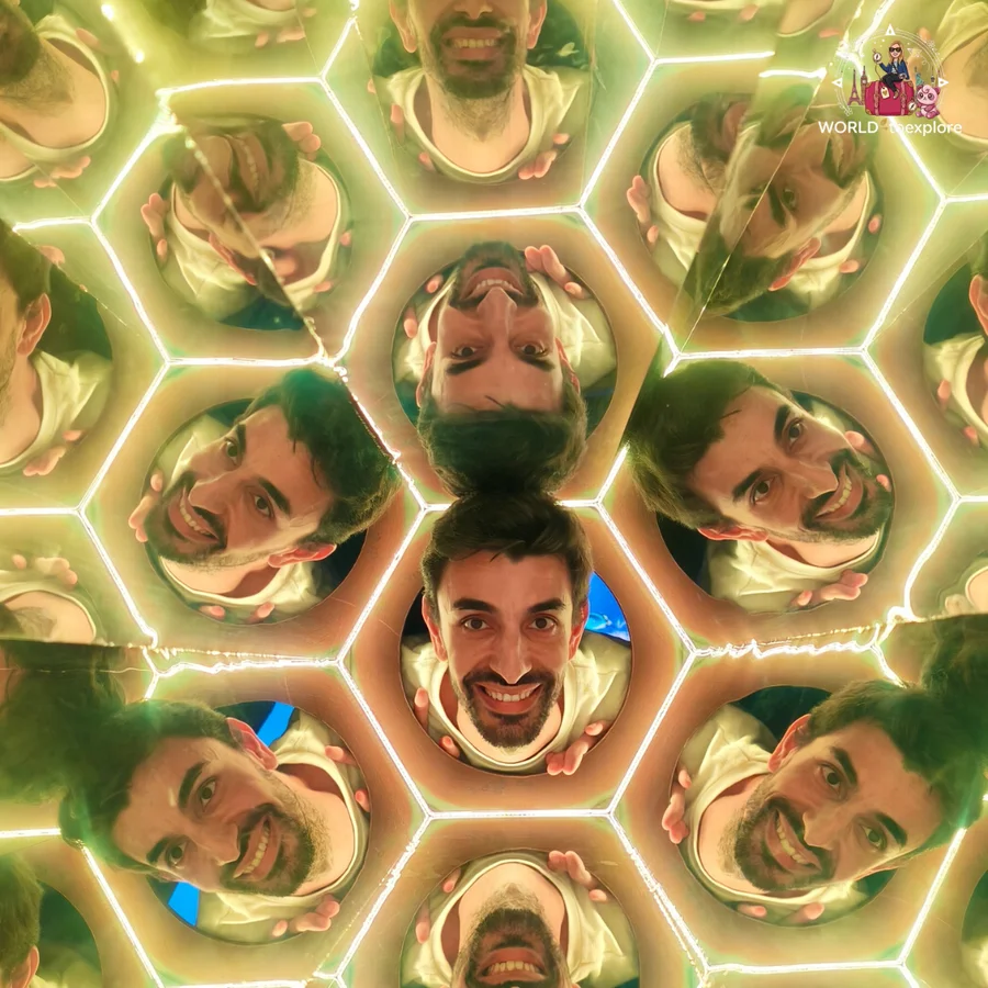 Her husband smiling in the middle of a hexagon mirror that multiplies his face, in a golden-lit room at Neonia