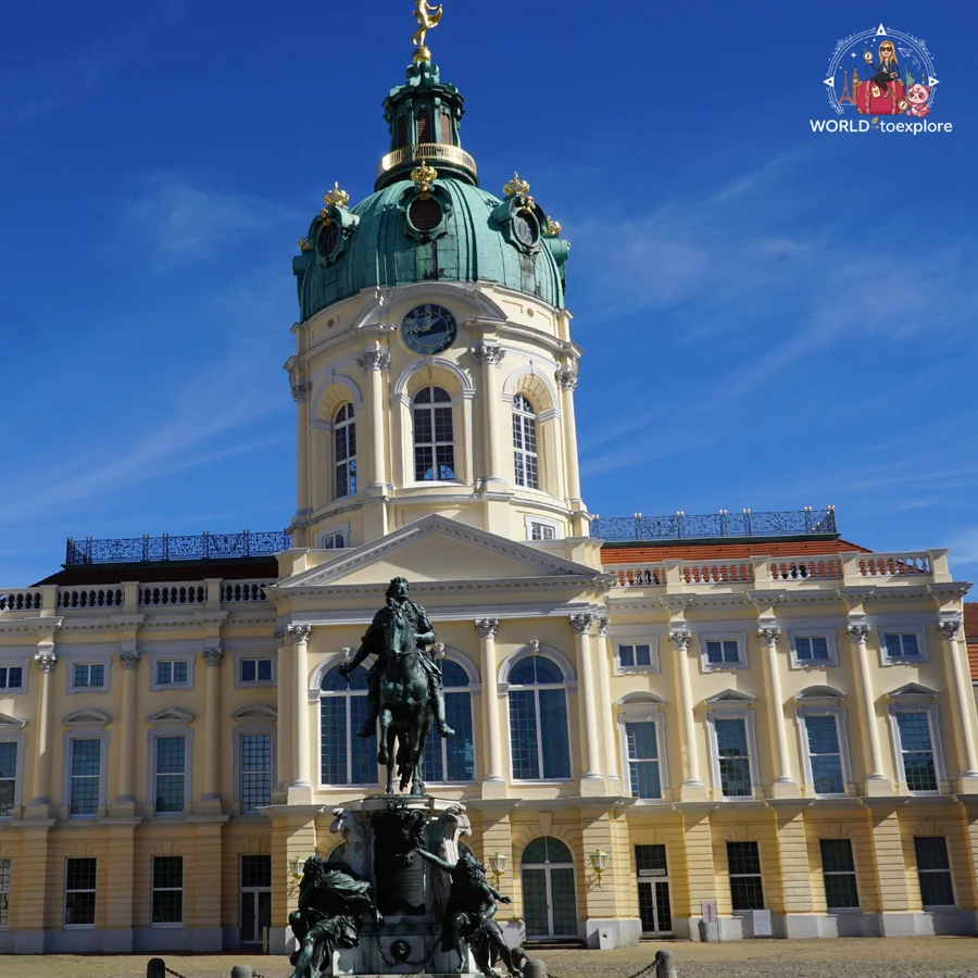Front façade of Charlottenburg Palace with its green dome and the equestrian statue of the Great Elector in the foreground, under a blue sky