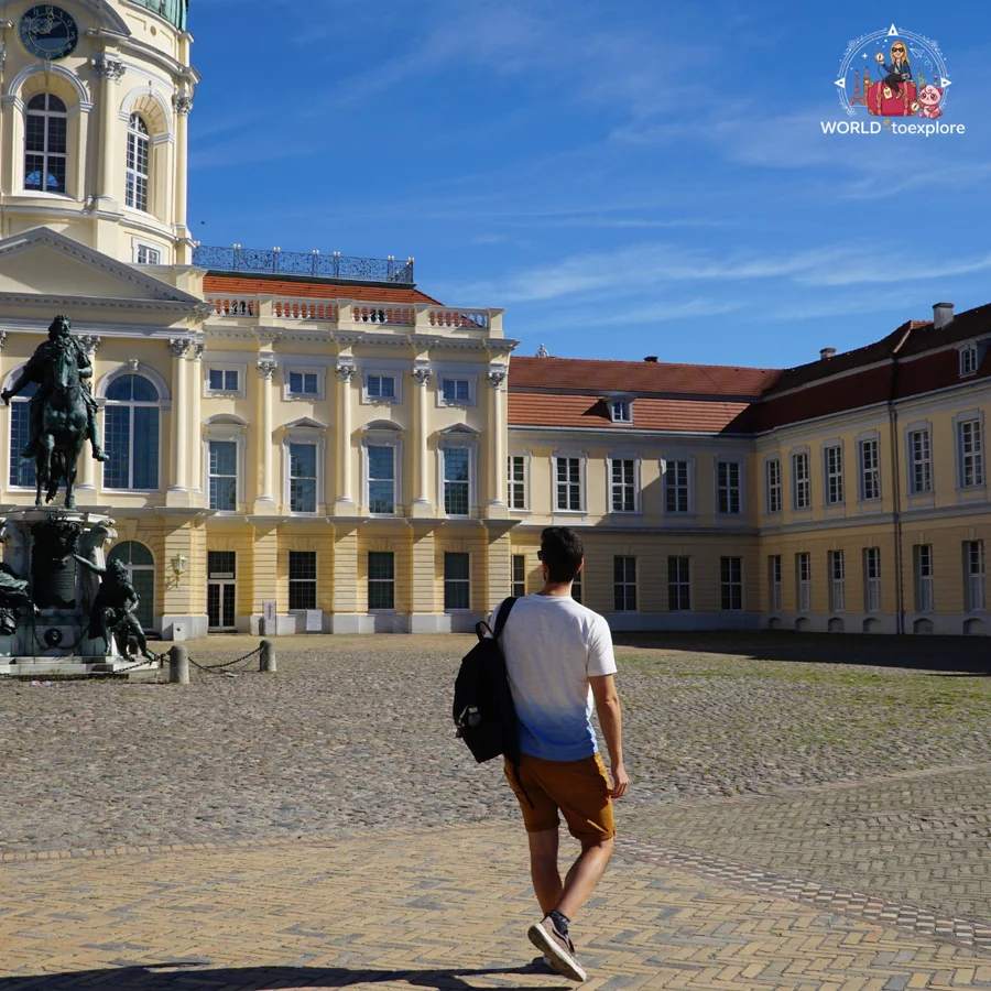 Man seen from behind, with a backpack, crossing the cobbled courtyard next to the equestrian statue and the palace's yellow wing