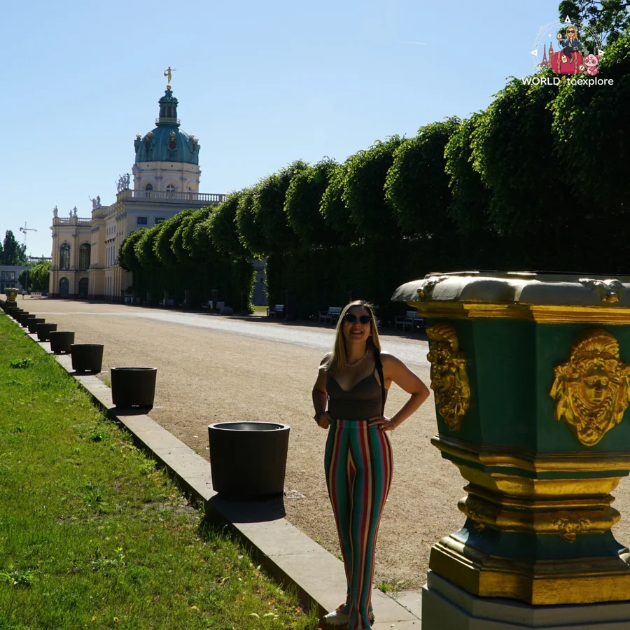 Woman standing on a gravel path next to a row of trimmed trees, with the palace in the background and a large green and gold vase in the foreground