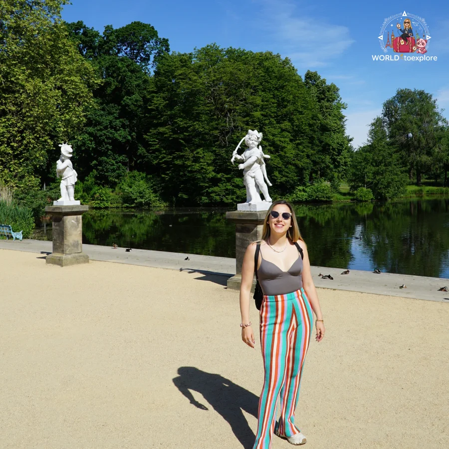 Woman walking along a gravel promenade by the lake, with two white statues and trees behind her