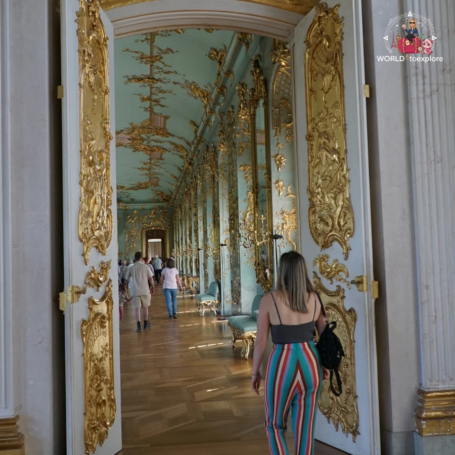 Woman seen from behind in a long gallery with white and gold doors and mirrors, with visitors in the background