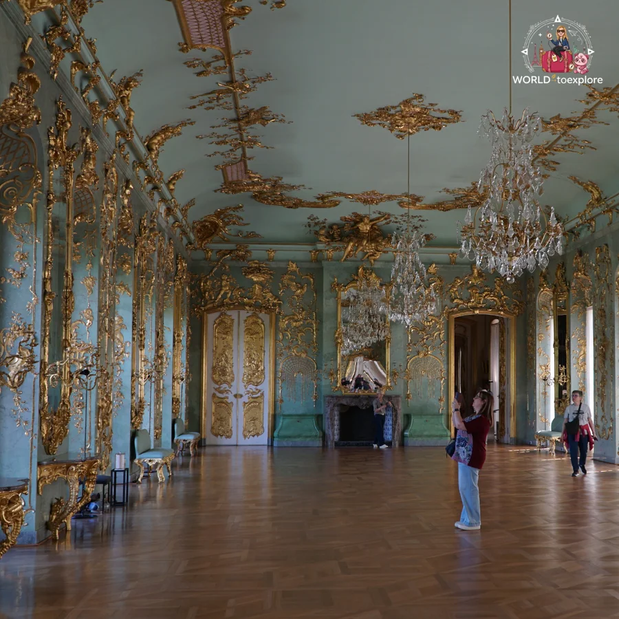 High-ceilinged room with aqua-green walls and gold ornaments, crystal chandeliers and two visitors in the background