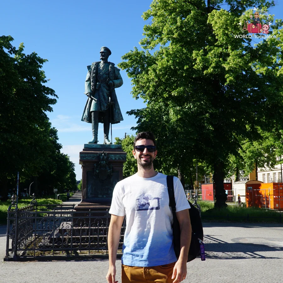 Man in sunglasses with a backpack next to a statue on a pedestal, under trees
