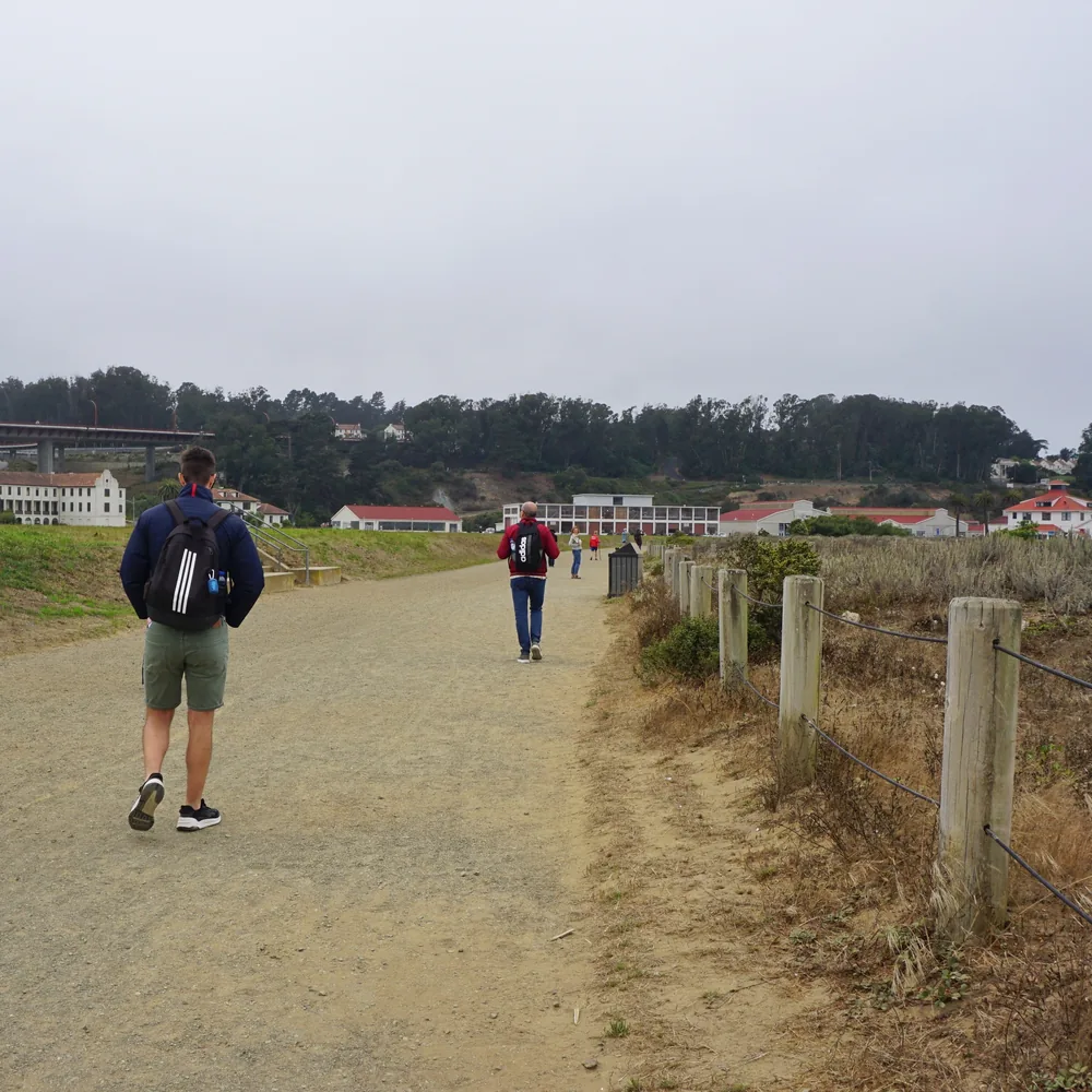 Two people walking along a dirt path at Crissy Field, with old red-roofed buildings and tree-covered hills in the background