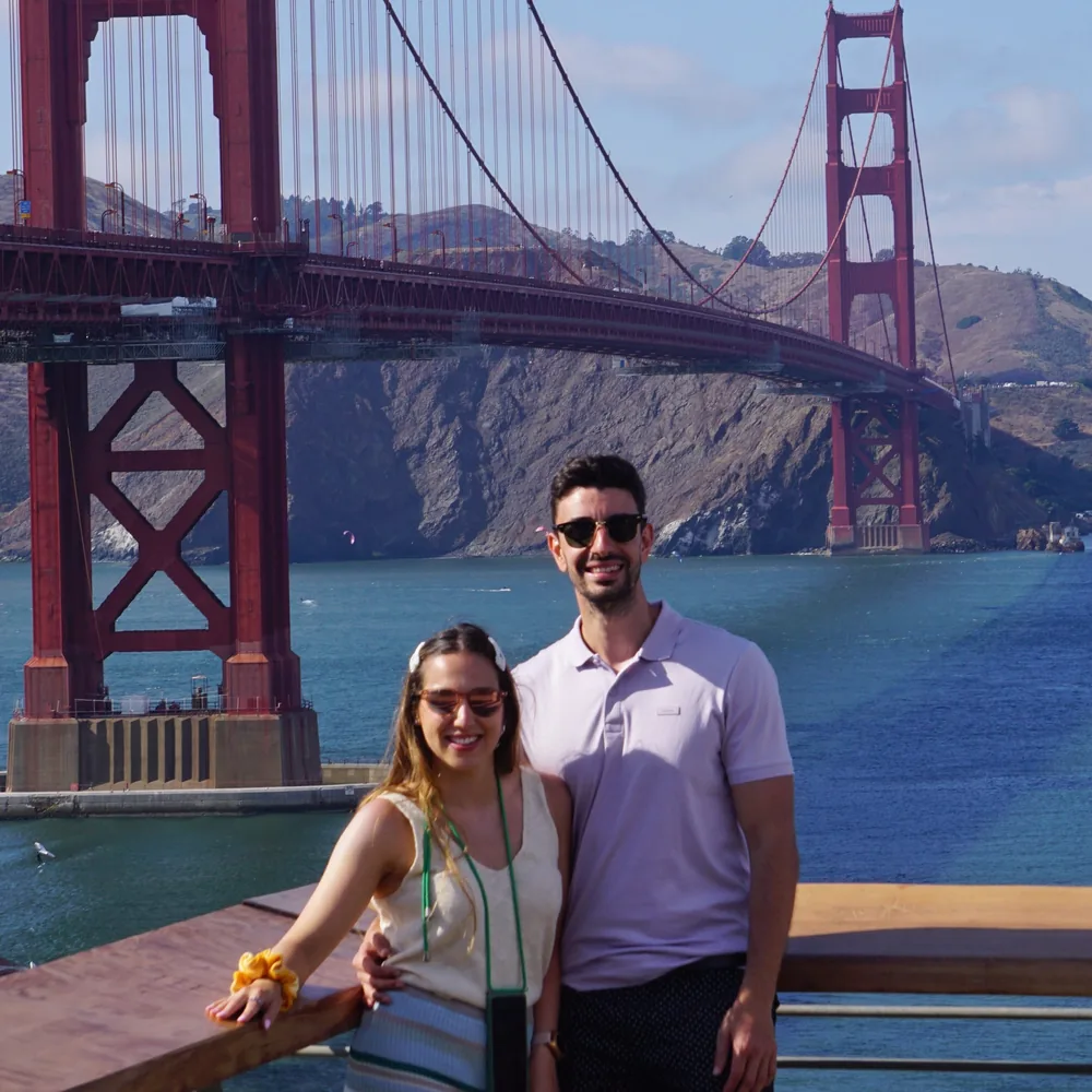 A couple posing at the Golden Gate Postcard Viewpoint, with the Golden Gate Bridge and the bay's hills in the background on a sunny day