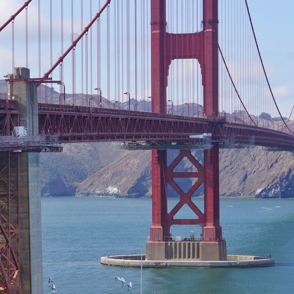 A close-up view of one of the Golden Gate Bridge's towers and its X-shaped bracing, with the bay and coastal hills behind