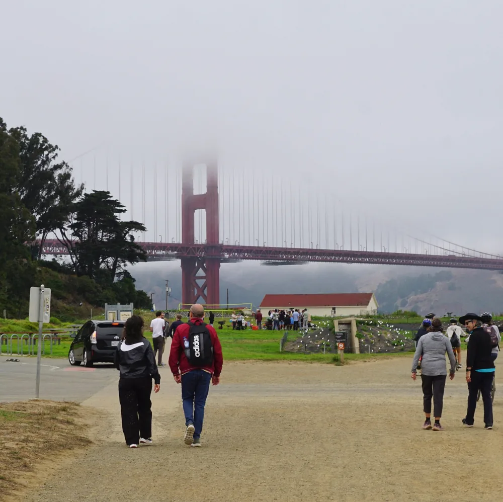 People walking towards the Golden Gate Bridge, partly hidden by fog, next to a grassy area at Crissy Field