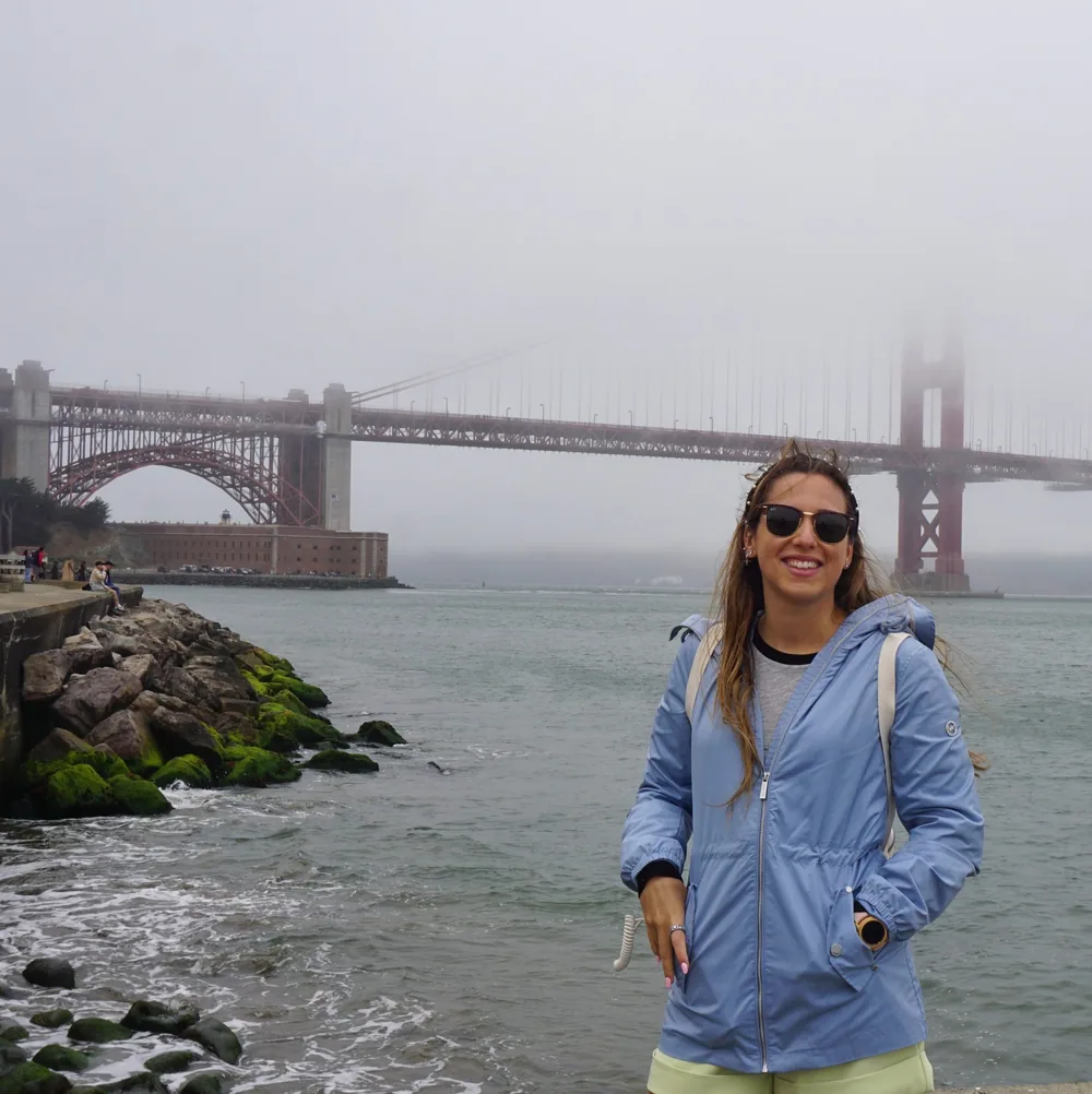 The author smiling by the rocky shoreline at the Golden Gate Viewpoint, with the fog-covered Golden Gate Bridge and Fort Point behind her