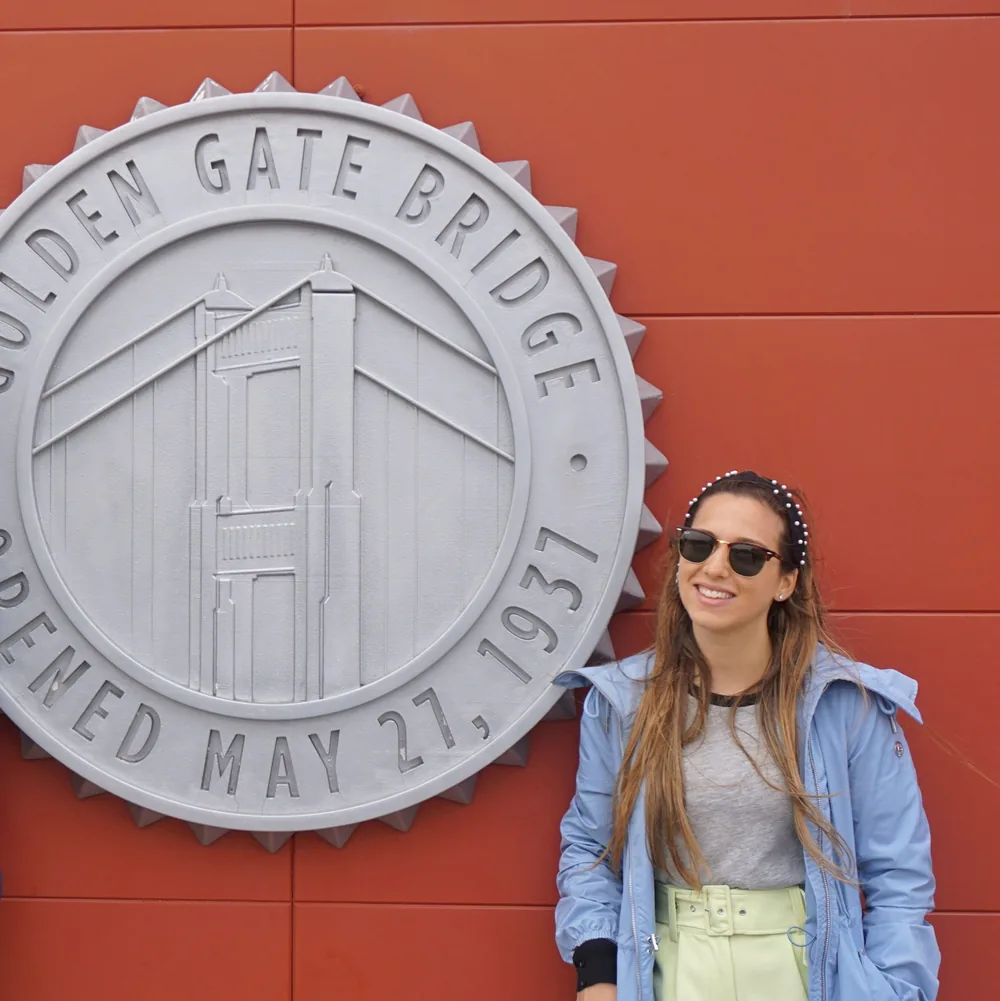 The author next to a circular metal plaque reading Golden Gate Bridge, Opened May 27, 1937, on the orange facade of the Welcome Center