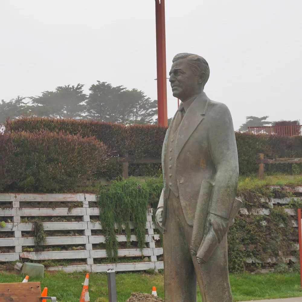 The bronze statue of Joseph Strauss, in a suit and tie, next to a garden with shrubs and a wooden fence