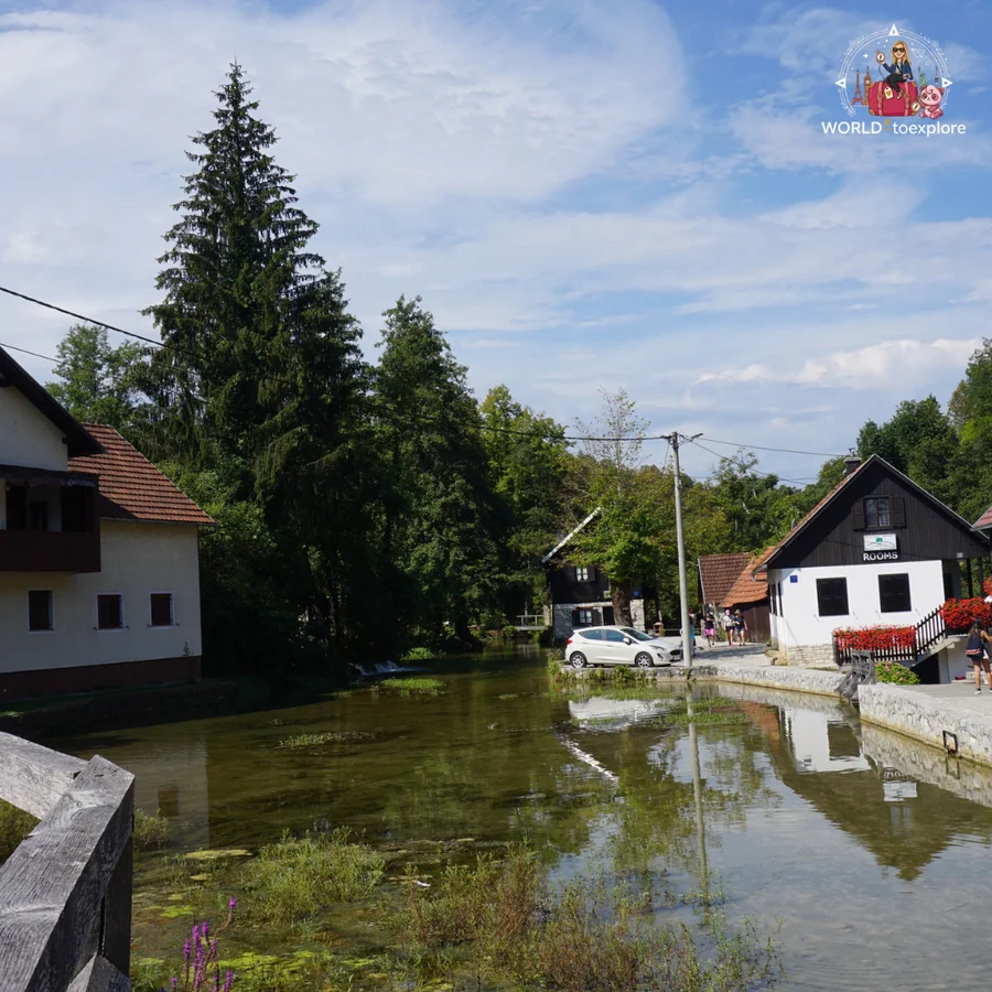 Channel of the Slunjčica river between the houses of Rastoke, with a white car by the water and a white house with a 'Rooms' sign