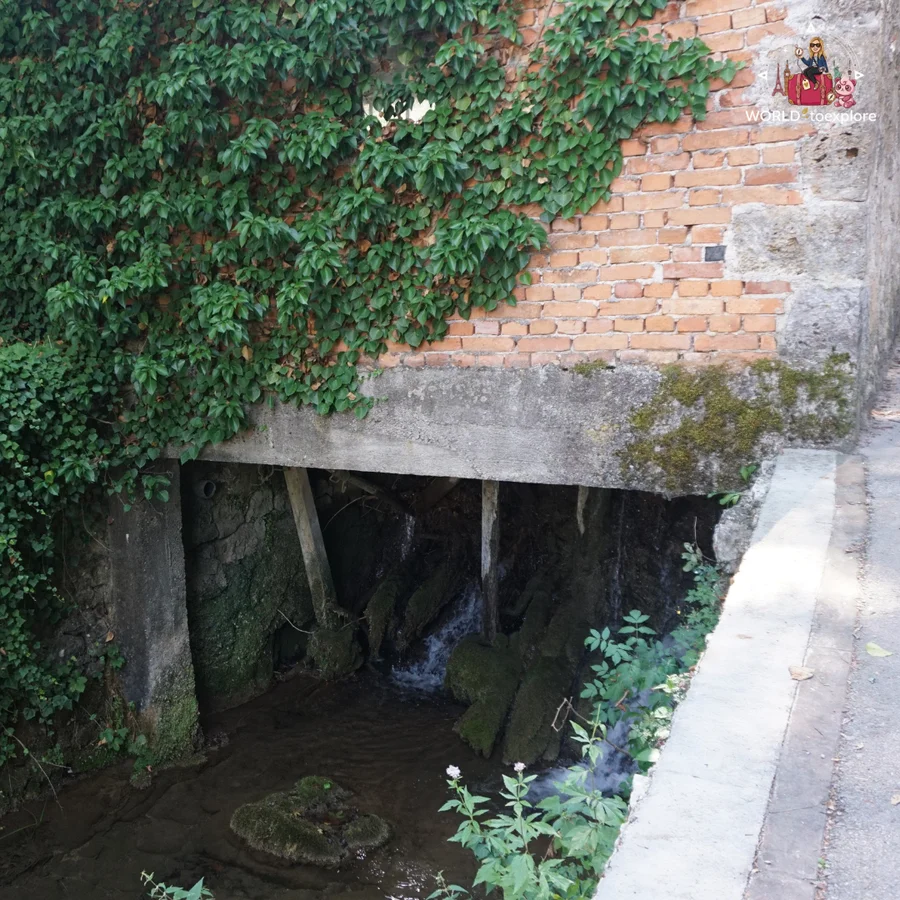 Water running under a house in Rastoke, between wooden posts and moss-covered stones, next to a path