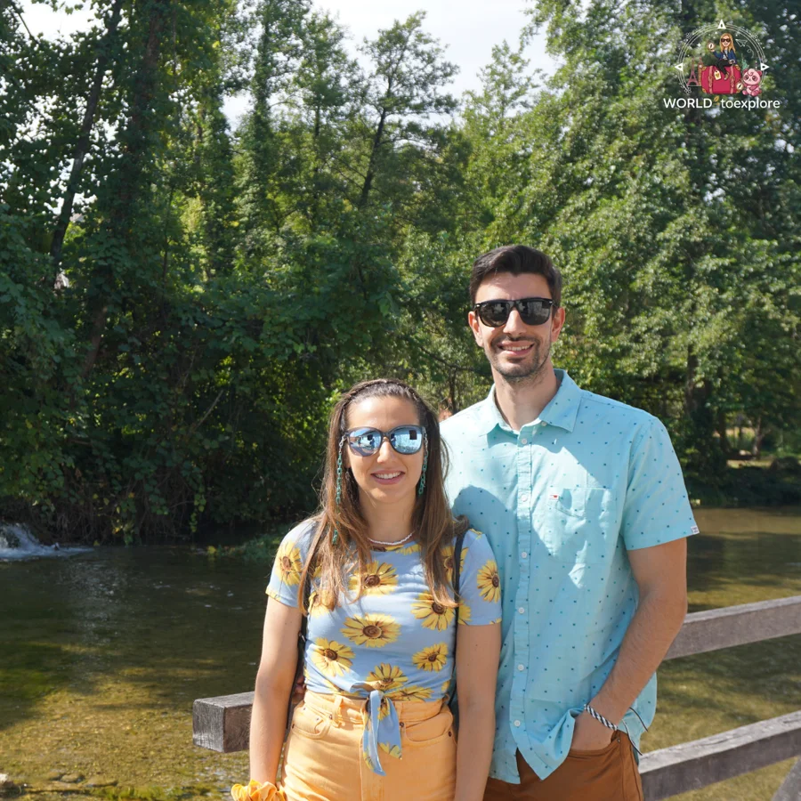 Cristina and her partner by the river in Rastoke, with green vegetation and a waterfall behind