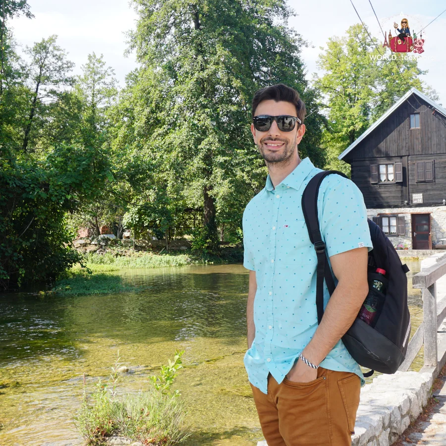Cristina's partner, with a backpack on, beside the clear river in Rastoke, with a dark wooden house behind