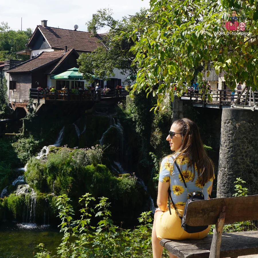 Cristina sitting on a wooden bench, looking at the waterfalls of Rastoke and the houses with terraces above