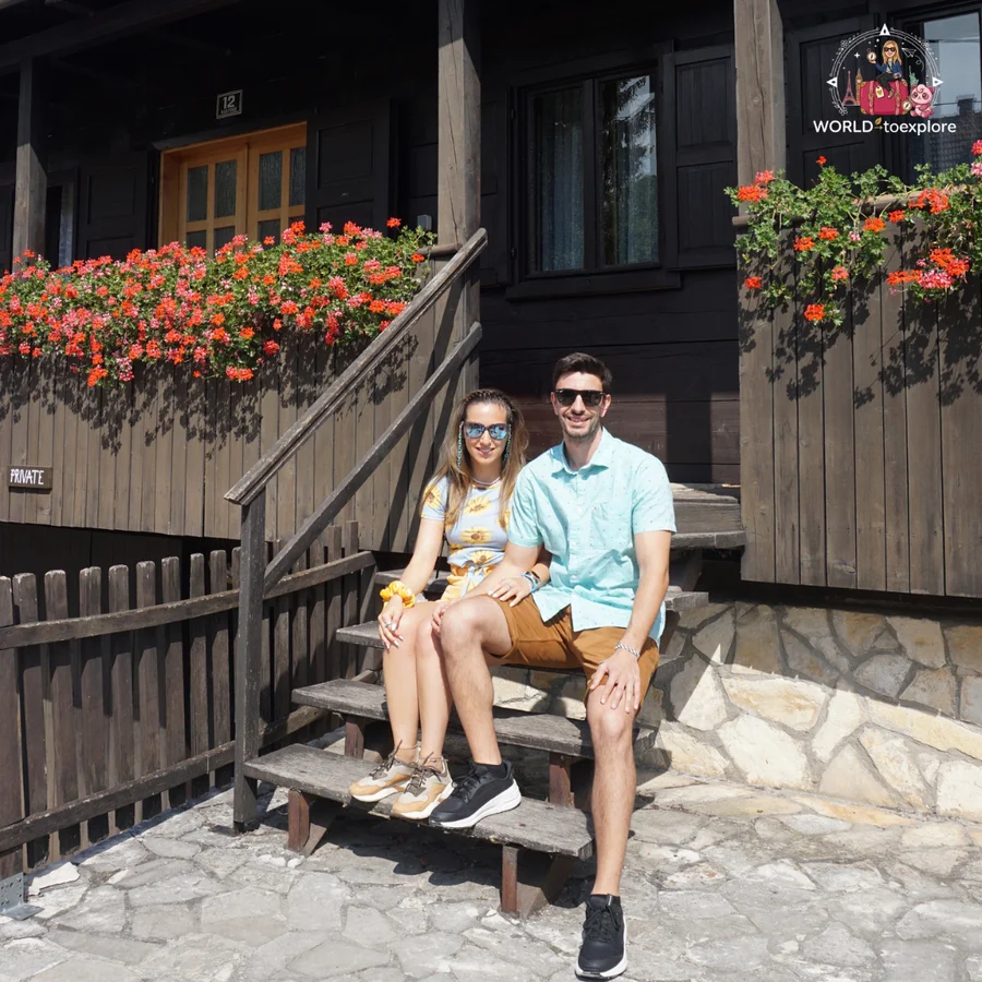 Cristina and her partner sitting on the wooden steps of a traditional Rastoke house, surrounded by red geraniums