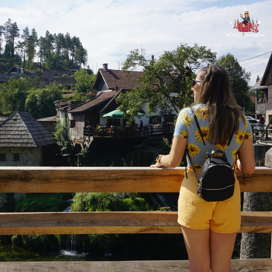 Cristina from behind, leaning on the wooden railing of the bridge, looking at the waterfalls and the houses of Rastoke