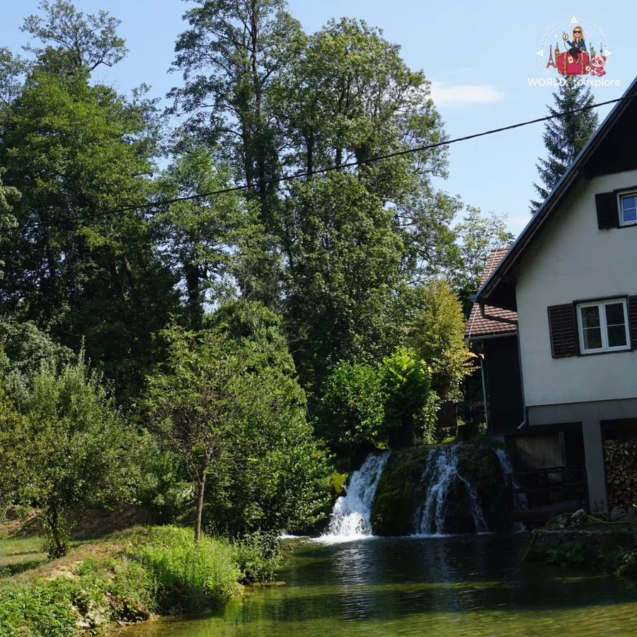 Waterfall tumbling beside a house in Rastoke into a pool of dark water, with stacked firewood on the right