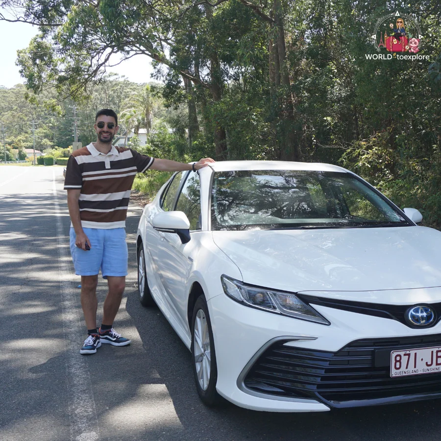 Man in a striped T-shirt next to a white Toyota sedan, on a road lined with trees