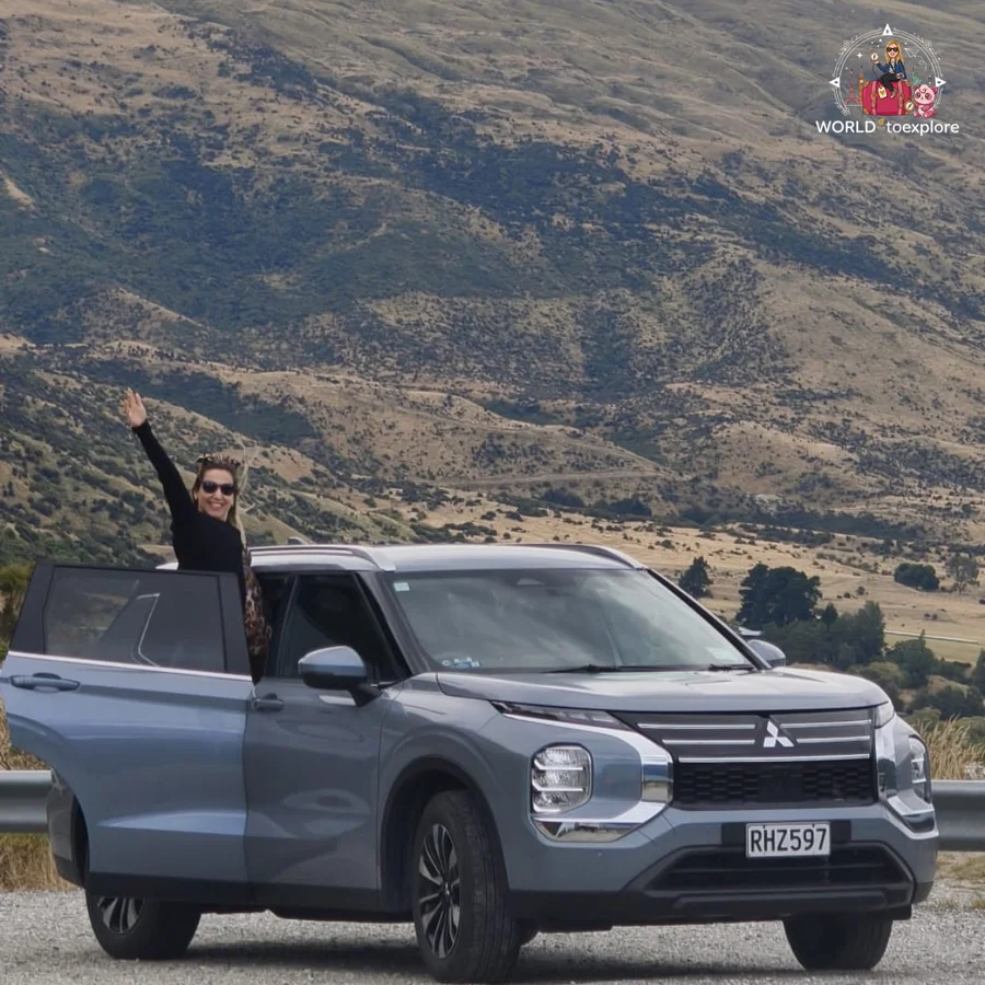 Woman waving from the open door of a grey Mitsubishi SUV, with golden hills behind