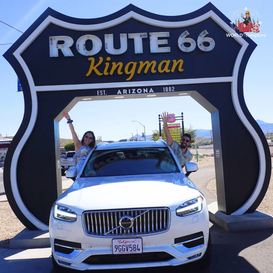 Couple waving from behind a white Volvo, inside the Route 66 Kingman sign arch in Arizona