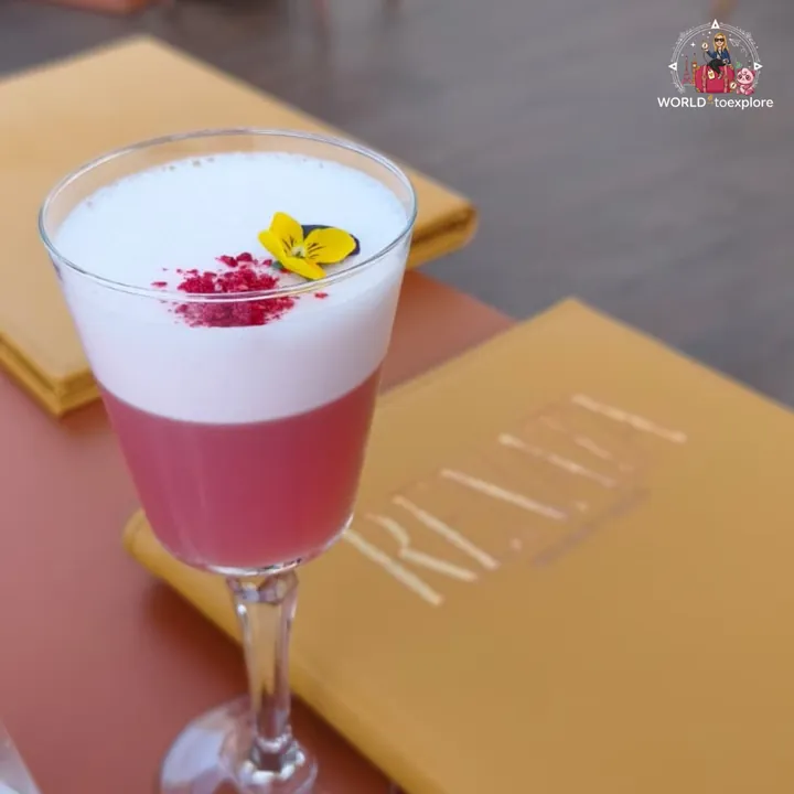 RENATA's signature cocktail, pink with white foam, an edible flower and raspberry crumble, resting on the restaurant's gold menu
