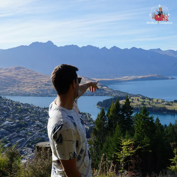 A man pointing at the view over Queenstown and Lake Wakatipu, with the mountains in the background