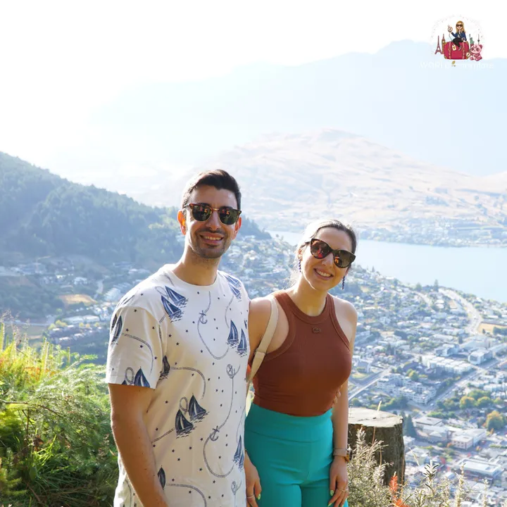 A smiling couple next to a tree stump, with Lake Wakatipu and Queenstown blurred in the background