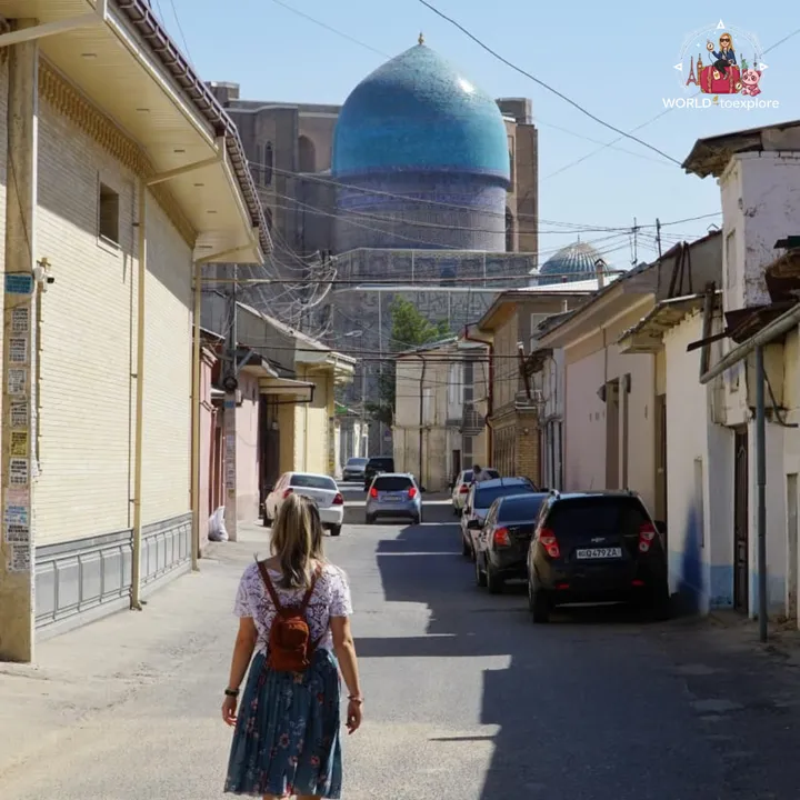 Woman walking down an old town street, with a large turquoise dome in the background