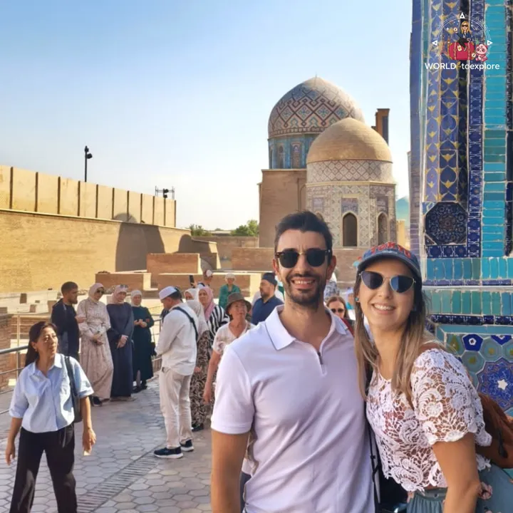 Smiling couple next to the ornate domes and blue tilework of a mausoleum complex, surrounded by other visitors