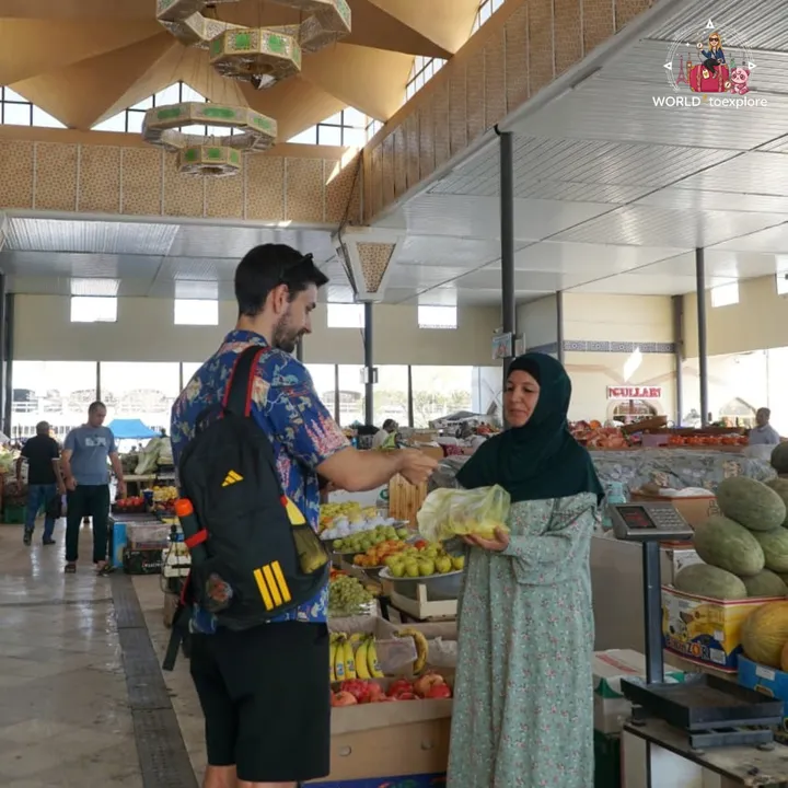 Man buying fruit from a headscarfed vendor at a covered market, among melons and other fresh produce