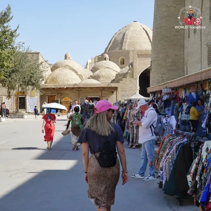 Woman walking through a market street lined with hanging traditional clothes, with historic domes in the background